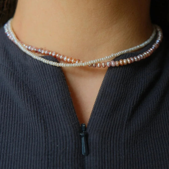 A close-up image of a neck wearing two elegantly intertwined pearl necklaces in shades of white and pink. The person is dressed in a textured dark top.
