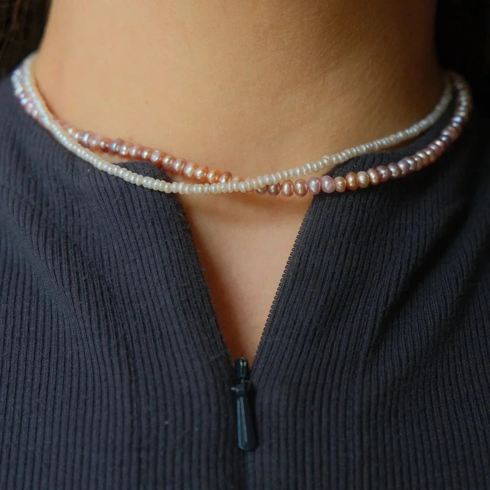 A close-up image of a neck wearing two elegantly intertwined pearl necklaces in shades of white and pink. The person is dressed in a textured dark top.