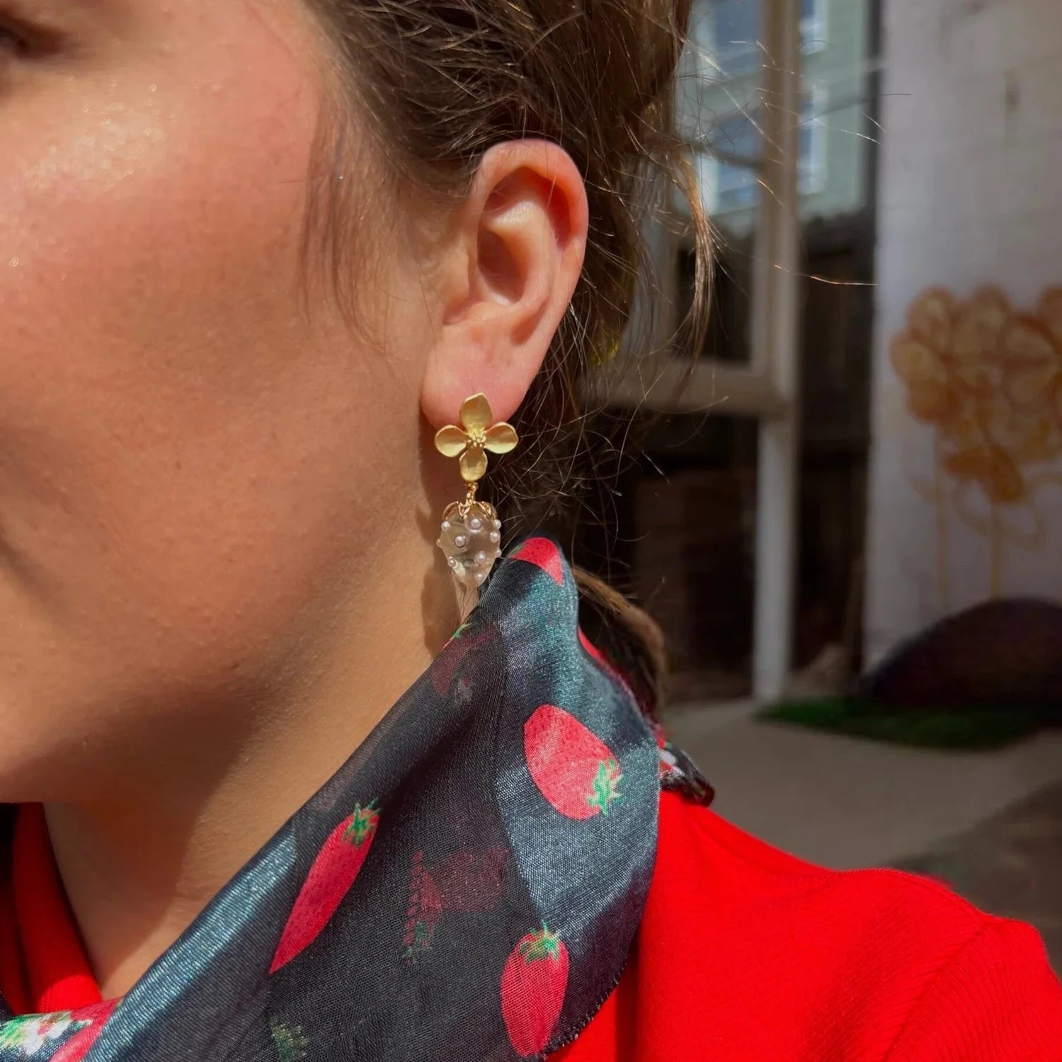 Close-up of a person wearing gold flower earrings with a drop clear and pearl strawberry, red jacket, and black scarf with strawberry pattern.