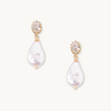Gold earrings with teardrop-shaped white pearls hanging from oval, faceted stones. The design is elegant and sophisticated on a plain background.