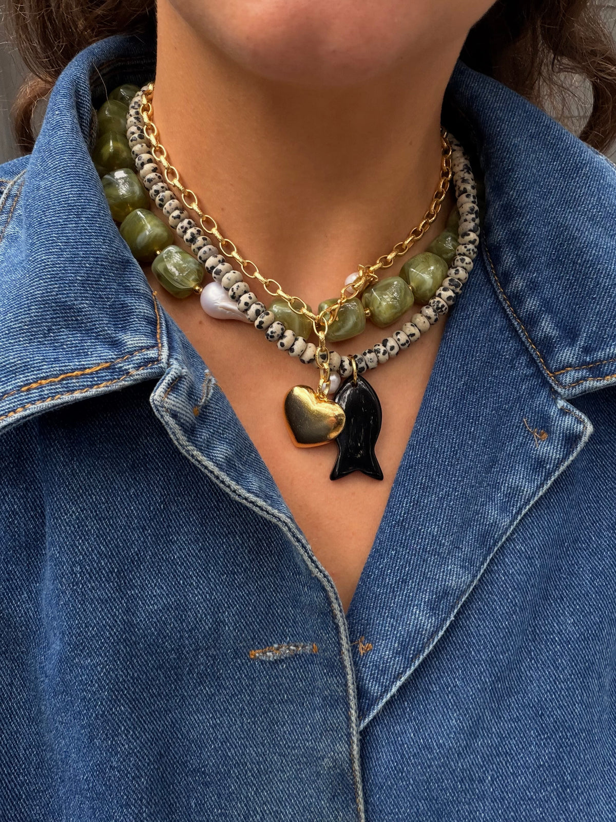 Woman wearing a denim jacket with a colorful fall necklace stack featuring green beads, jasper Dalmatian beads and a gold heart necklace. 