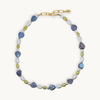 A pearl necklace featuring heart-shaped iridescent blue and purple, white baroque pearls, olive green beads, and a gold-tone chain clasp, displayed on a light cream background.