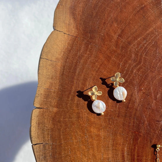 Gold-tone flower earrings with pearl drops on a wooden surface. The sunlight casts soft shadows, creating a warm and elegant atmosphere.