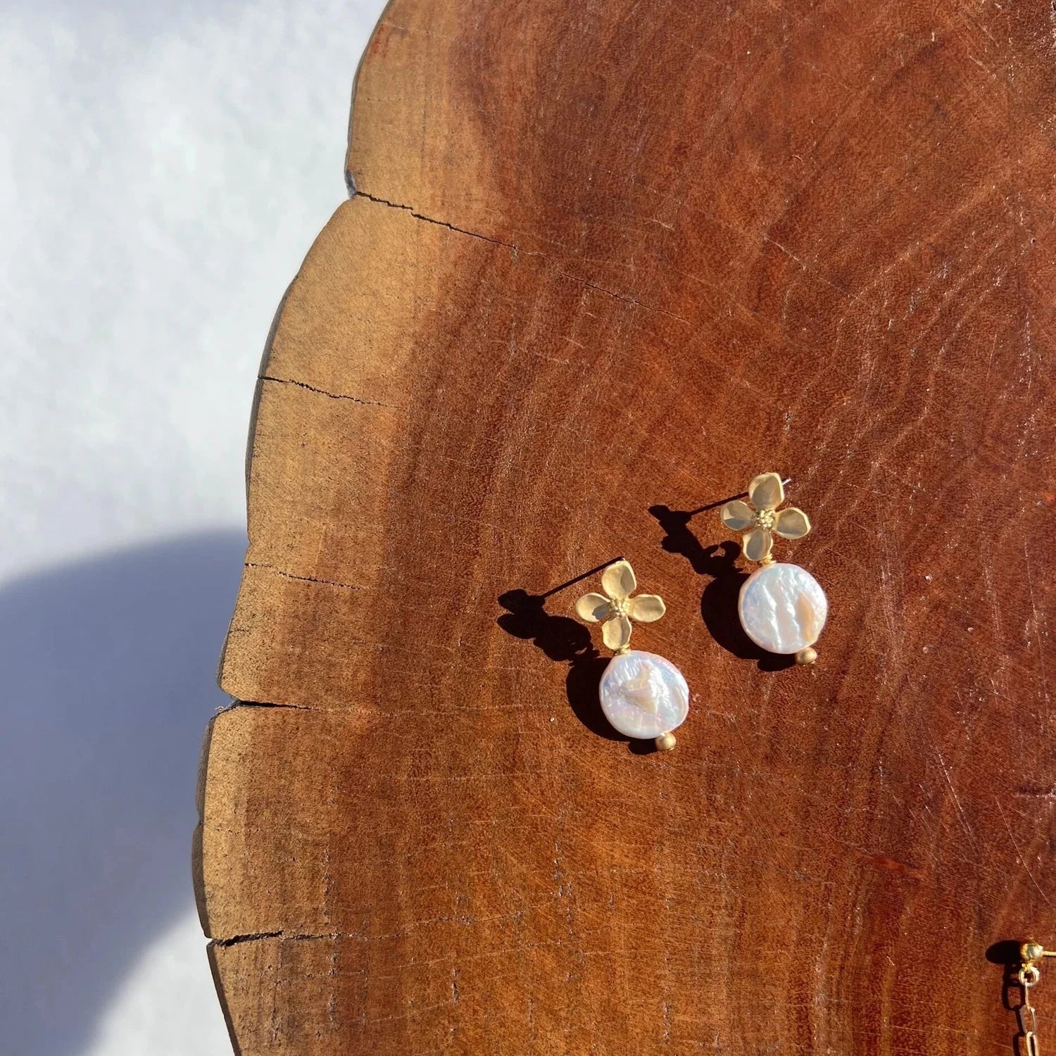 Gold-tone flower earrings with pearl drops on a wooden surface. The sunlight casts soft shadows, creating a warm and elegant atmosphere.