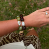 A close-up of a person's wrist wearing two beaded bracelets outdoors on a grassy lawn. One bracelet features irregular white mother-of-pearl beads, and the other features dark brown and olive green beads.