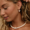 A close-up lifestyle shot of a woman with blonde hair wearing a white beaded necklace and a small gold hoop earring with a white heart-shaped charm.