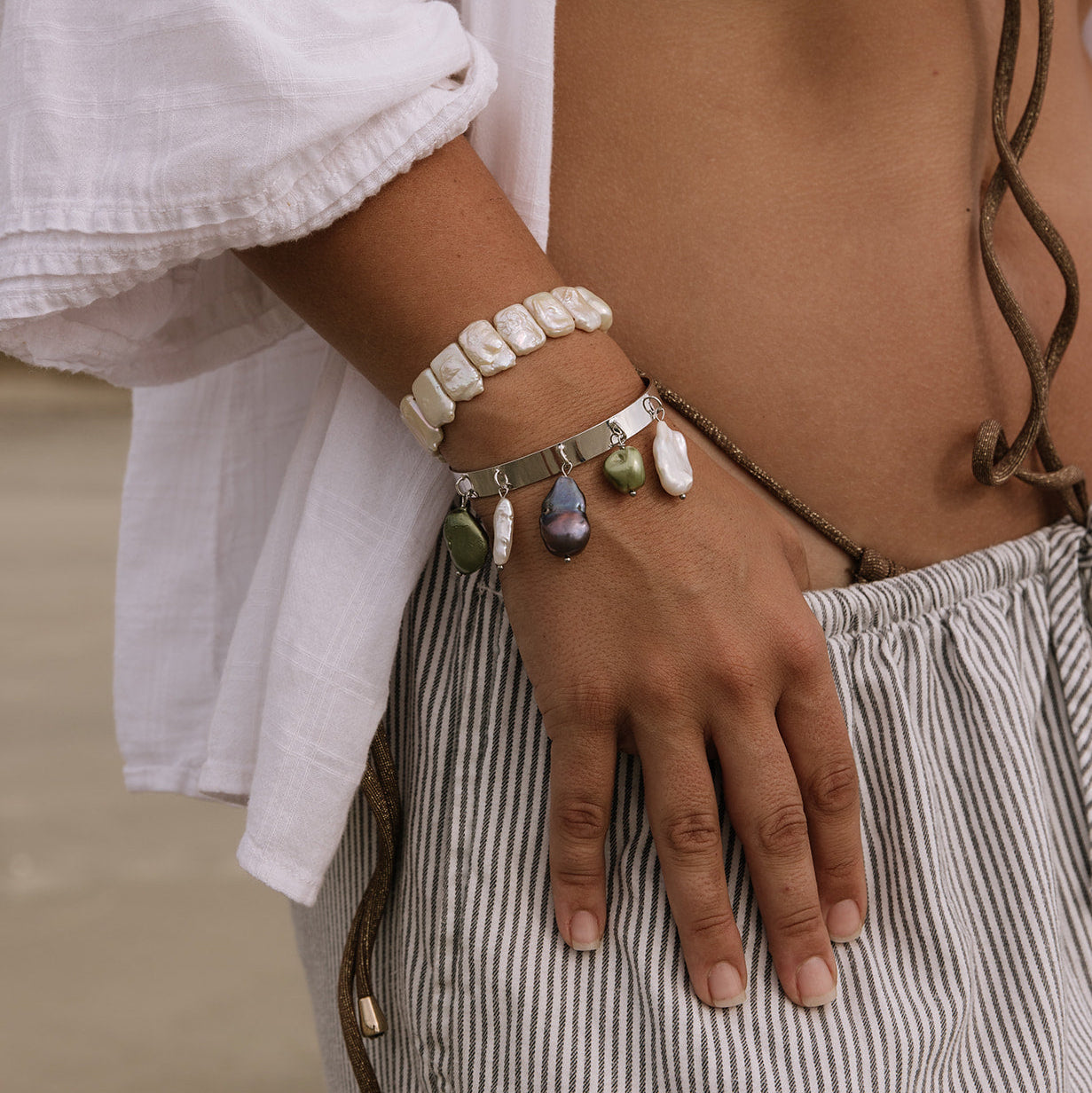 A close-up of a person's hand and midsection wearing multiple bohemian-style bracelets, including a white beaded bracelet, a silver-toned chain bracelet with colorful stone charms, and a dark cord bracelet.