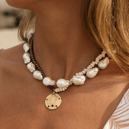 A close-up of a woman's neck and chest wearing a bohemian-style double-strand necklace. The necklace features large, irregular white baroque pearls, small cream-colored seed beads, a dark brown cord, and a gold-toned starfish pendant.