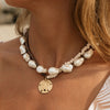 A close-up of a woman's neck and chest wearing a bohemian-style double-strand necklace. The necklace features large, irregular white baroque pearls, small cream-colored seed beads, a dark brown cord, and a gold-toned starfish pendant.