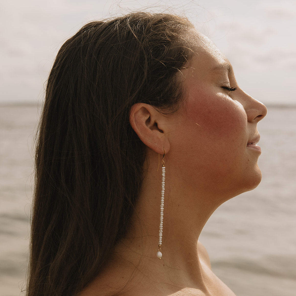 A woman in profile wearing a long, dangling silver beaded earring, with a soft-focus beach or water background.