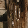 Two bohemian-style necklaces hanging from a weathered wooden post. One necklace features a gold chain with a stacked stone pendant.