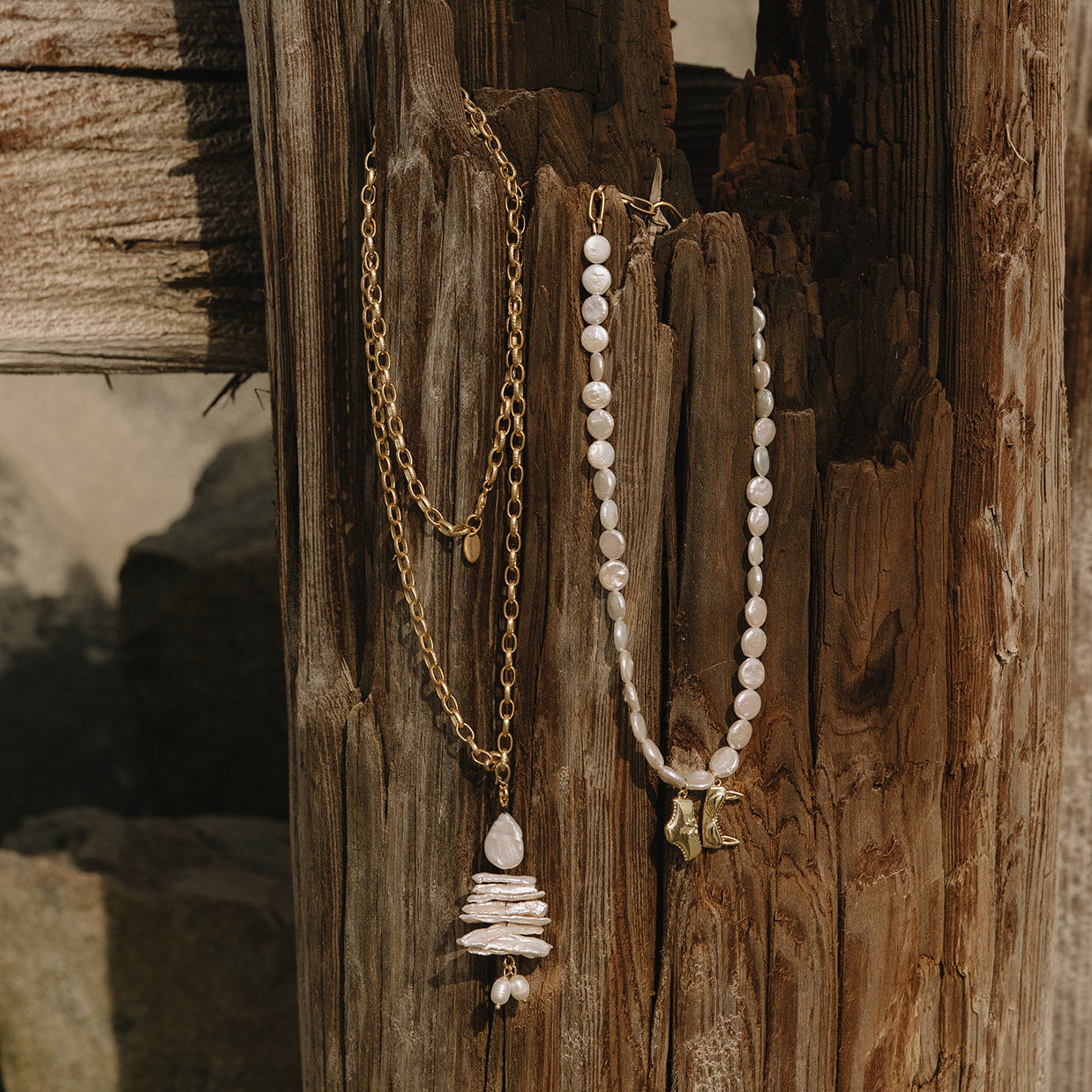 Two bohemian-style necklaces hanging from a weathered wooden post. One necklace features a gold chain with a stacked stone pendant.