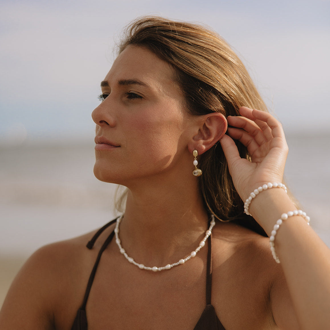 A woman on a beach wearing a white shell-bead necklace, matching shell-bead bracelets, and dangling shell earrings, wearing a dark brown bikini top.