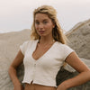 A woman posing on a sandy beach wearing a cream-colored, short-sleeve knit crop top with a button-down front and black bikini bottoms.