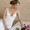 Woman in a white satin slip dress holding a bouquet of flowers on a bed wearing gold and pearl vintage inspired jewelry