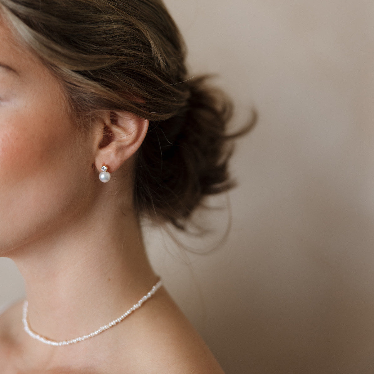 Side profile of a woman with wavy hair in a bun, wearing a pearl earring and delicate necklace, against a soft, neutral background. Elegant and serene mood.