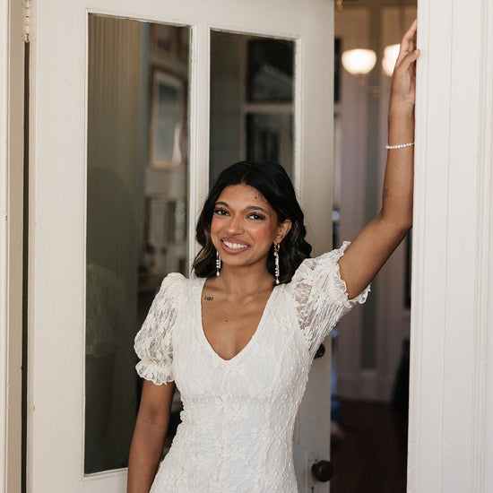 A smiling woman in a white lace dress stands in a doorway, arm resting on the frame. She wears dangling earrings, exuding elegance and joy.