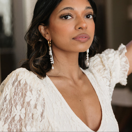 Woman in a lace white dress and long earrings gazes confidently. Her expression is serene, with soft makeup and dark wavy hair framing her face.