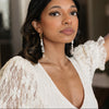 Woman in a lace white dress and long earrings gazes confidently. Her expression is serene, with soft makeup and dark wavy hair framing her face.