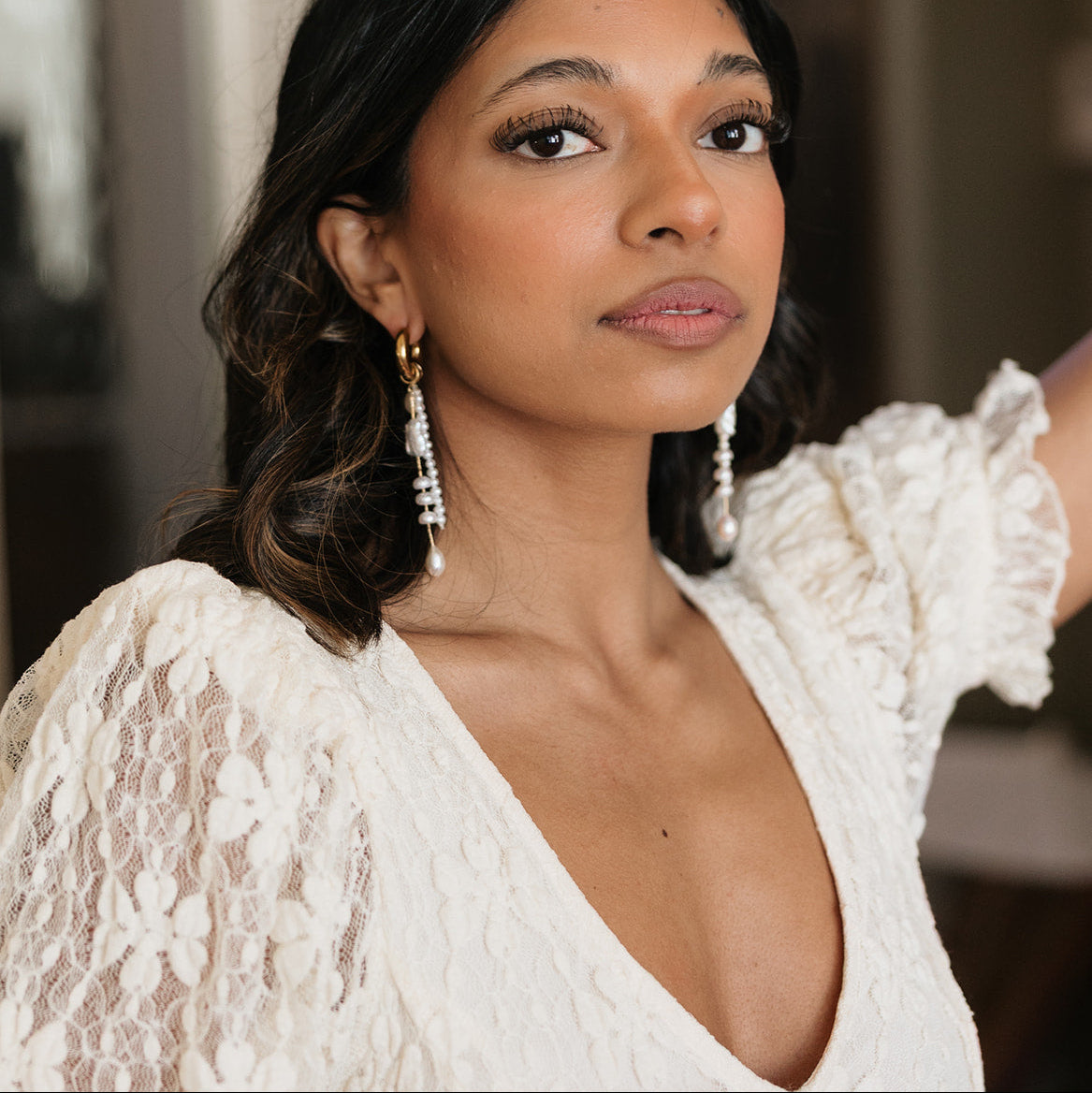 Woman in a lace white dress and long earrings gazes confidently. Her expression is serene, with soft makeup and dark wavy hair framing her face.
