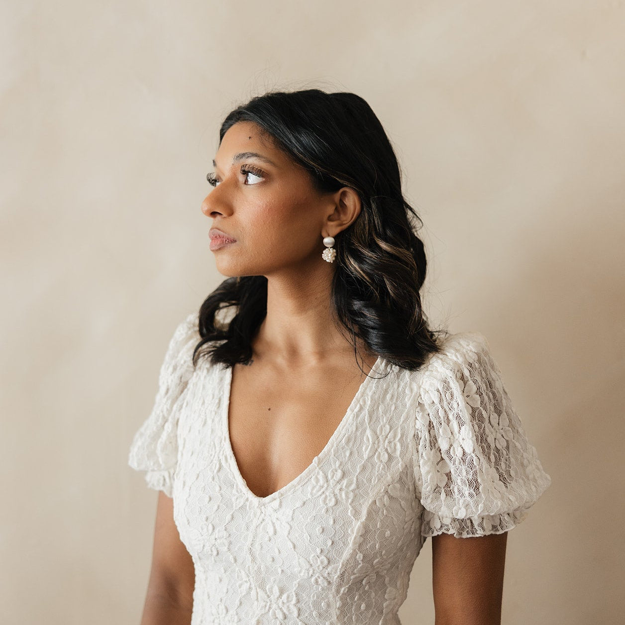 A woman in a white lace top gazes to the left, her expression calm and thoughtful. She has wavy hair and wears pearl drop earrings.