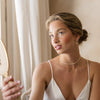 Woman in a white satin slip holding a hand mirror indoors wearing delicate pearl bridal jewelry.