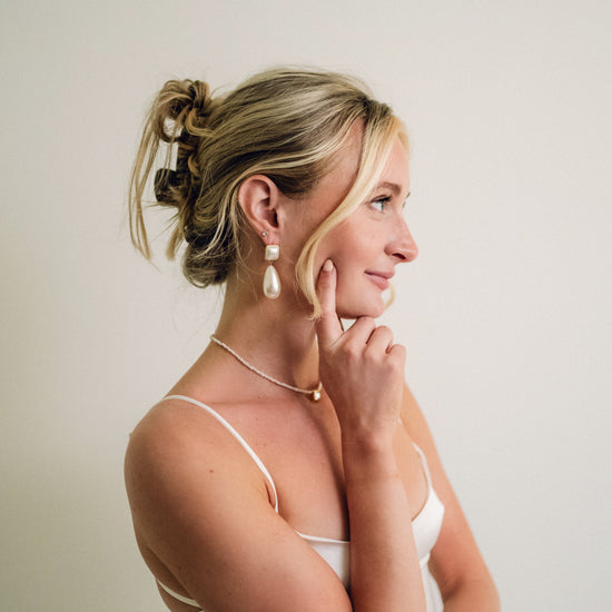 Woman in profile with blonde hair in a loose bun, wearing pearl earrings and necklace, touching her chin. The tone is elegant and contemplative.