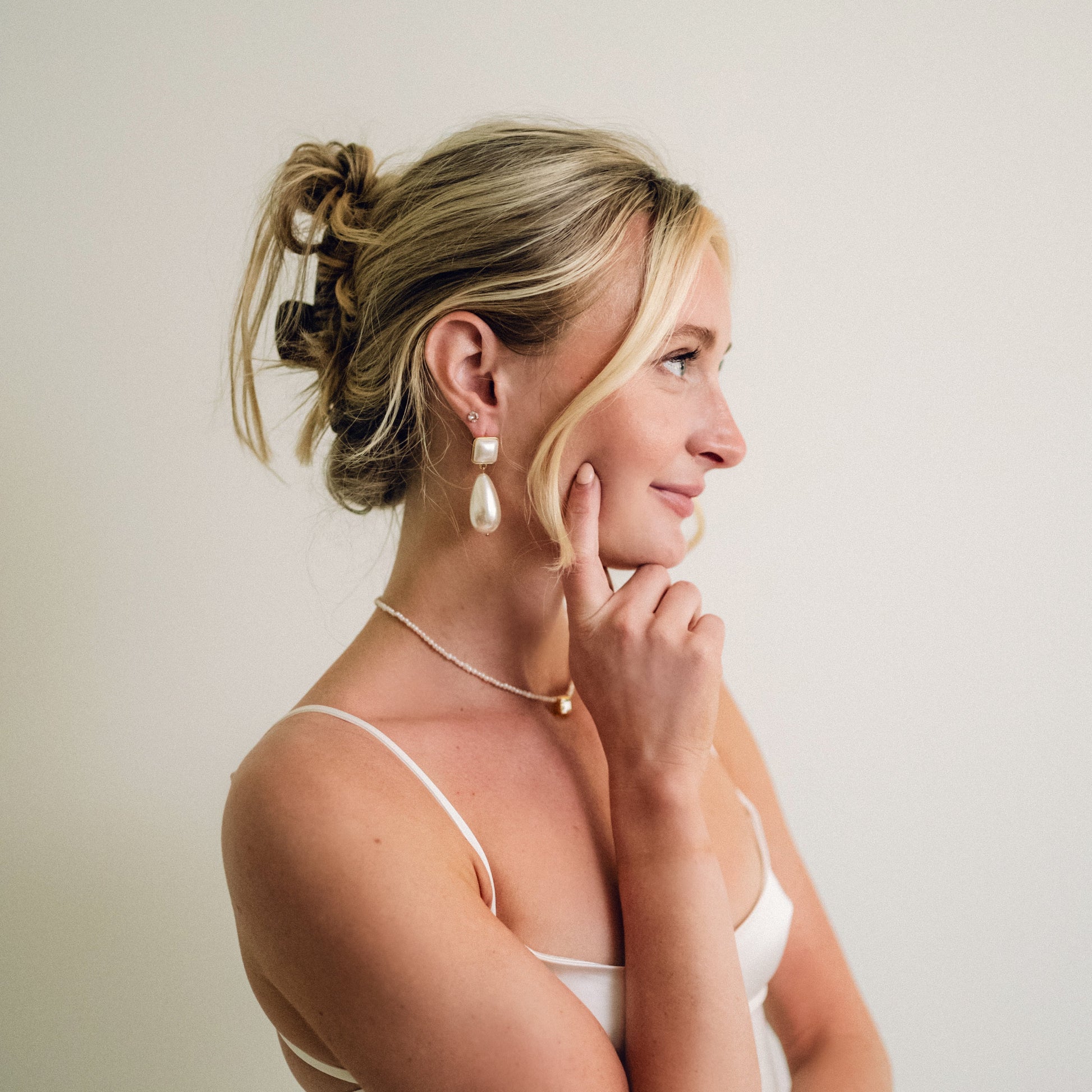 Woman in profile with blonde hair in a loose bun, wearing pearl earrings and necklace, touching her chin. The tone is elegant and contemplative.