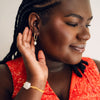 A woman with braided hair, wearing a red patterned top, gently touches an elegant pearl hoop earring. She has a serene expression, conveying calmness and confidence.