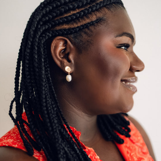 A woman with braided hair, wearing a red patterned top, gently touches an elegant earring. She has a smiling expression, conveying calmness and confidence.