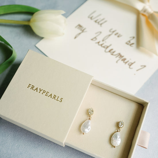 A pair of pearl earrings in a FRAYPEARLS box rests on a gray surface. Nearby, a card asks "Will you be my bridesmaid?" alongside a white tulip. Elegant and inviting.