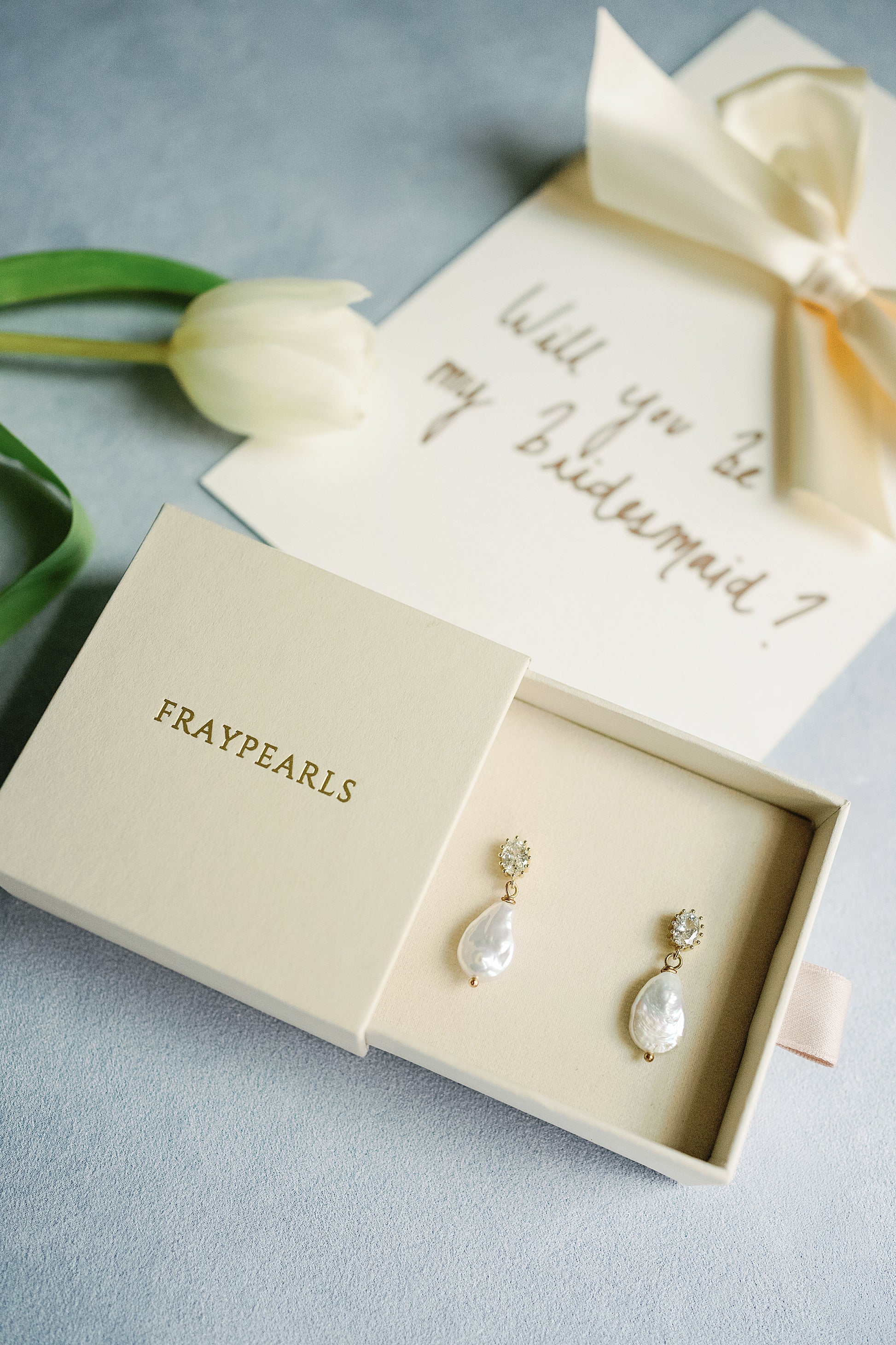 A pair of pearl earrings in a FRAYPEARLS box rests on a gray surface. Nearby, a card asks "Will you be my bridesmaid?" alongside a white tulip. Elegant and inviting.