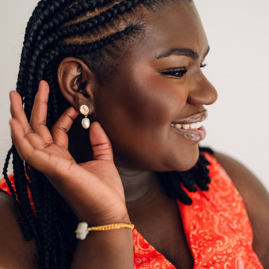 A woman with braided hair, wearing a red patterned top, gently touches an elegant earring. She has a smiling expression, conveying excitement and confidence.