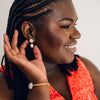 A woman with braided hair, wearing a red patterned top, gently touches an elegant earring. She has a smiling expression, conveying excitement and confidence.