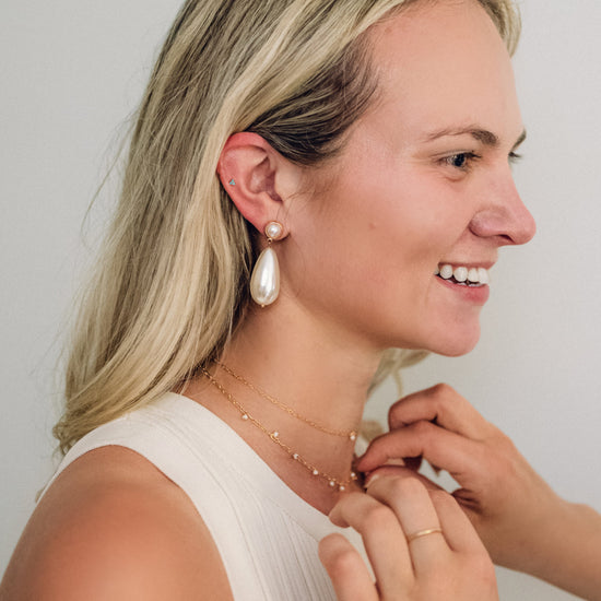 A woman with long blonde hair smiles, showcasing a large pearl earring. She touches delicate gold necklaces, exuding elegance and joy.