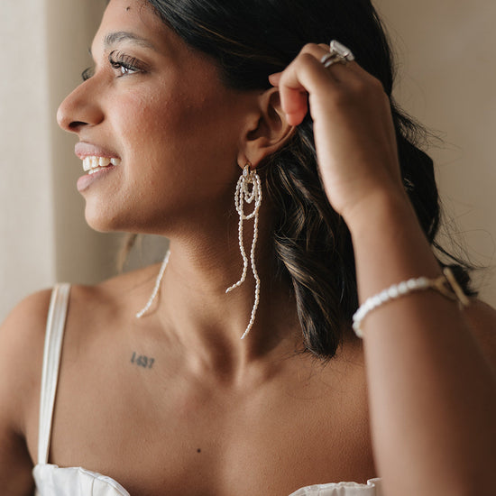 A woman smiles gently, gazing sideways. She wears long, elegant pearl earrings and a silver ring. Her dark hair contrasts with her light outfit.