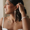 A woman smiles gently, gazing sideways. She wears long, elegant pearl earrings and a silver ring. Her dark hair contrasts with her light outfit.