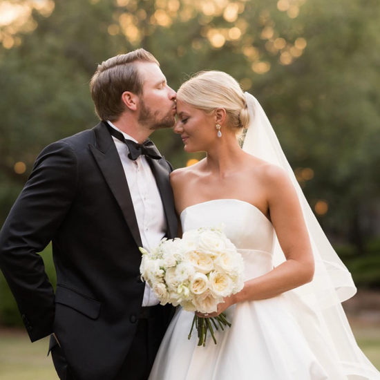 A groom in a tuxedo gently kisses the bride's forehead in a strapless gown, holding a bouquet of white roses; a serene and loving moment captured outdoors.
