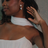 A woman wearing a white wrap around her shoulders, features a teardrop earring, with a serene expression and hand raised near her hair.
