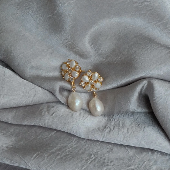 Elegant pearl earrings on a textured silver fabric. The earrings feature gold floral settings with teardrop pearls, conveying sophistication and luxury.