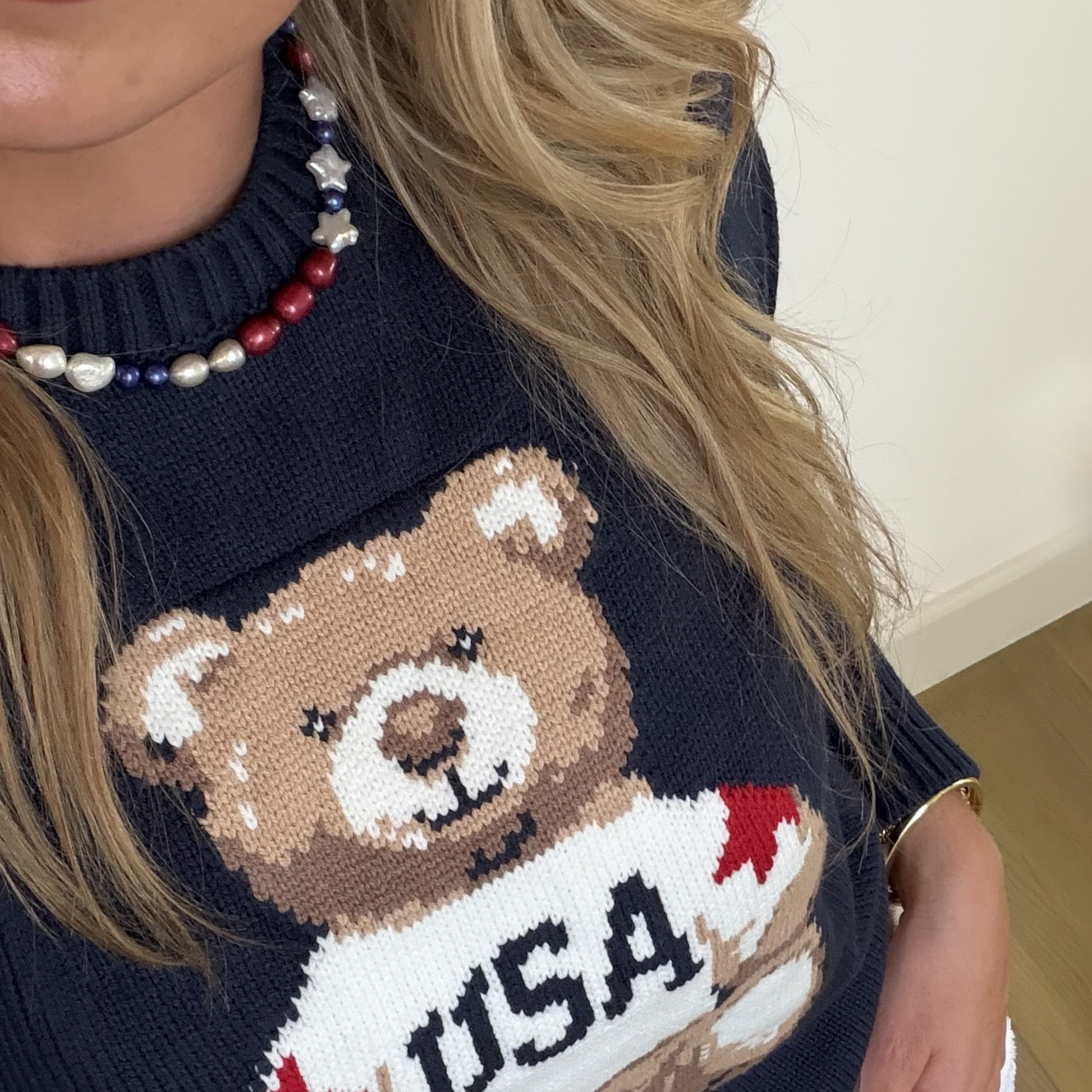 Close-up of a person wearing a navy sweater with a cute bear design. They have long, wavy blonde hair and a red, white, and blue pearl necklace.