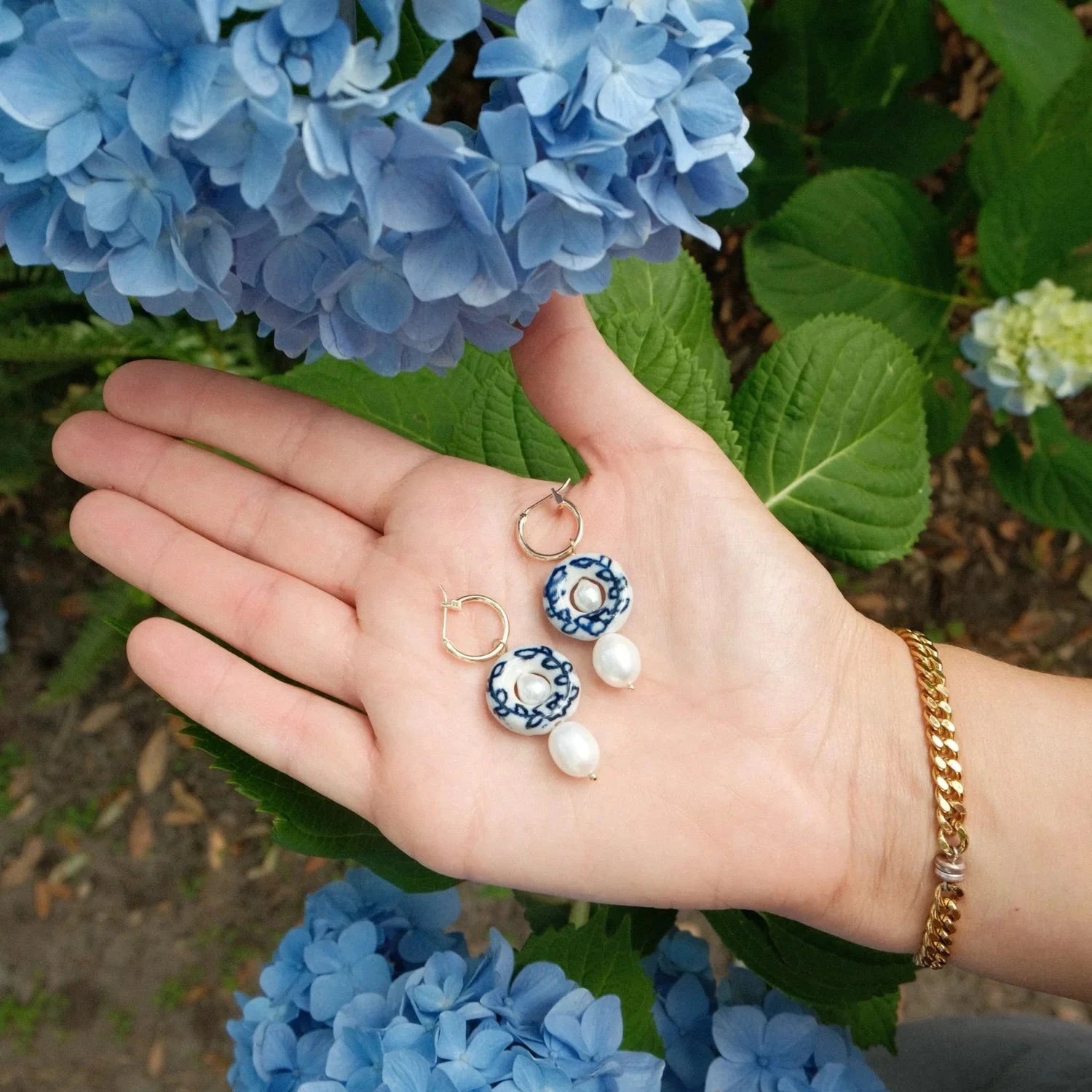 Hand holding a pair of earrings with blue and white floral designs, surrounded by blue flowers.