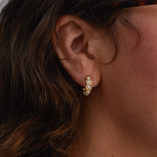 Close-up of a person's ear wearing a gold earring with three round pearls. The earring is elegant and eye-catching against their dark hair.