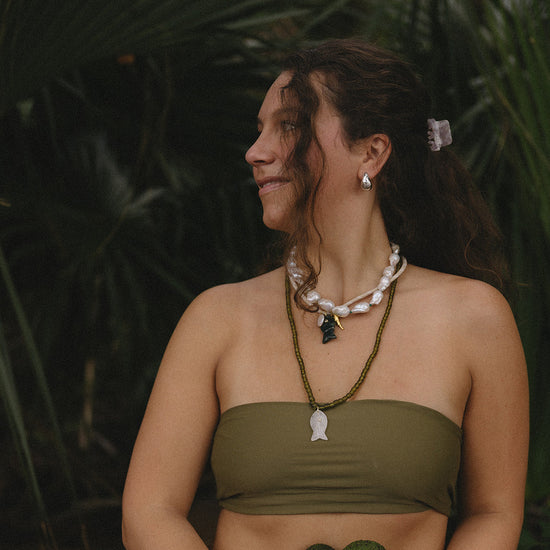 Woman with long curly hair wearing a green strapless top and layered necklaces, stands in profile against lush greenery; she looks content and serene.
