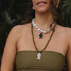 Woman with long curly hair wearing a green strapless top and layered necklaces, stands against lush greenery; she looks content and serene.