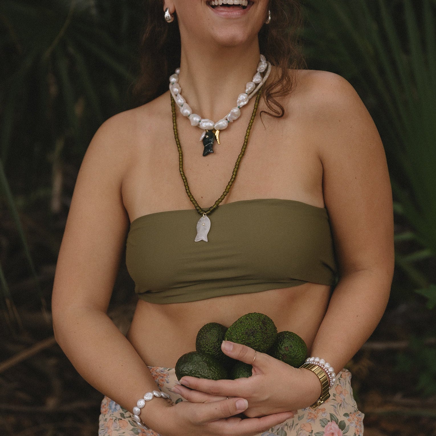 Woman with long curly hair wearing a green strapless top and layered necklaces, stands holding avocados against lush greenery; she looks content and serene.