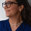 A smiling person with tortoiseshell glasses and wavy hair wears pearl earrings and a matching necklace, dressed in a blue scrub top, conveying a sense of elegance.