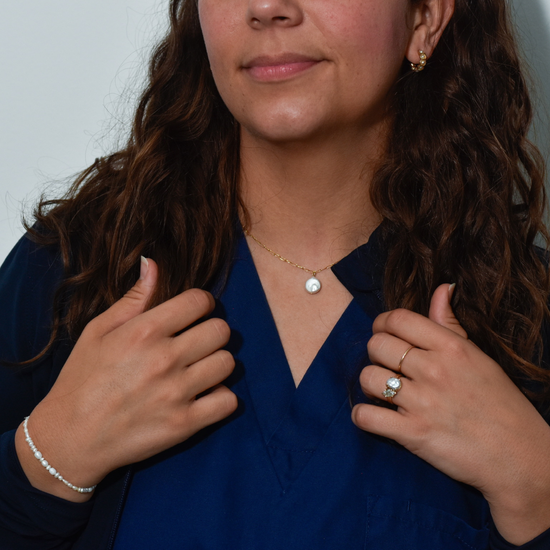 A person with wavy brown hair wearing a blue top is shown from the shoulders down. They have a delicate gold necklace with a pearl pendant, matching earrings, a pearl bracelet, and a ring adorned with a large pearl. The tone is calm and elegant.