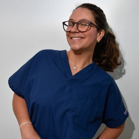 Woman in blue scrubs smiling warmly against a plain backdrop. She wears glasses, a heart necklace, and her hair is tied back, conveying a friendly tone.