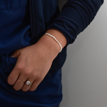 A close-up of a person in navy clothing with a hand in their pocket. They wear a delicate pearl bracelet and a ring with a large stone, illustrating elegance.